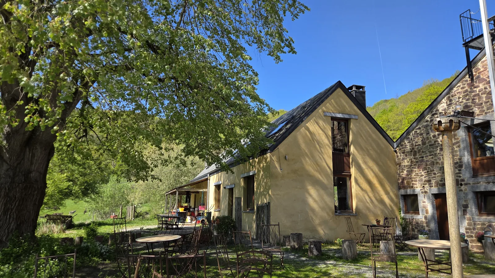La petite maison jaune des 4 Sources, sous un grand arbre, terrasse extérieure avec tables et chaises devant, ciel bleu, falaise boisée à l'arrière