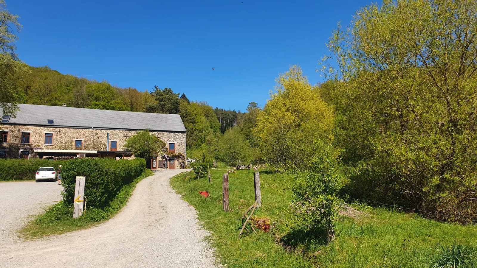 Chemin de gravier qui mène au bâtiment principal en pierre, terrasse à l'avant, voiture à gauche, haie de buissons et arbres en pleine pousse de printemps, ciel bleu