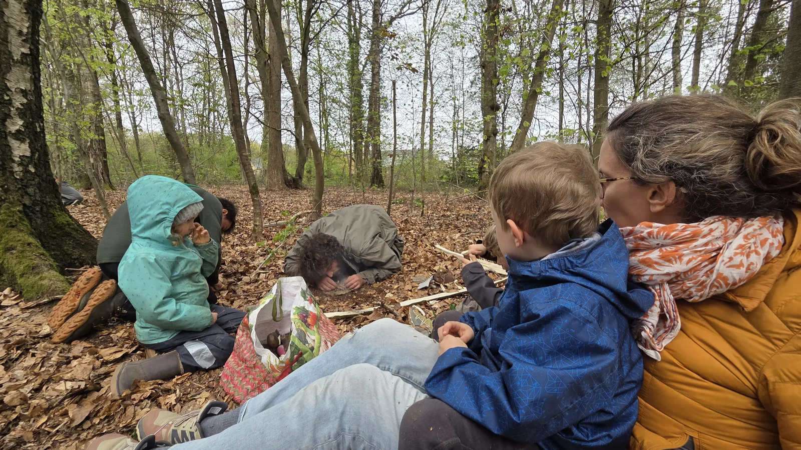 Plusieurs enfants et adultes assis dans les feuilles mortes en sous-bois, penchés ensemble vers le sol pour suivre l'allumage du feu
