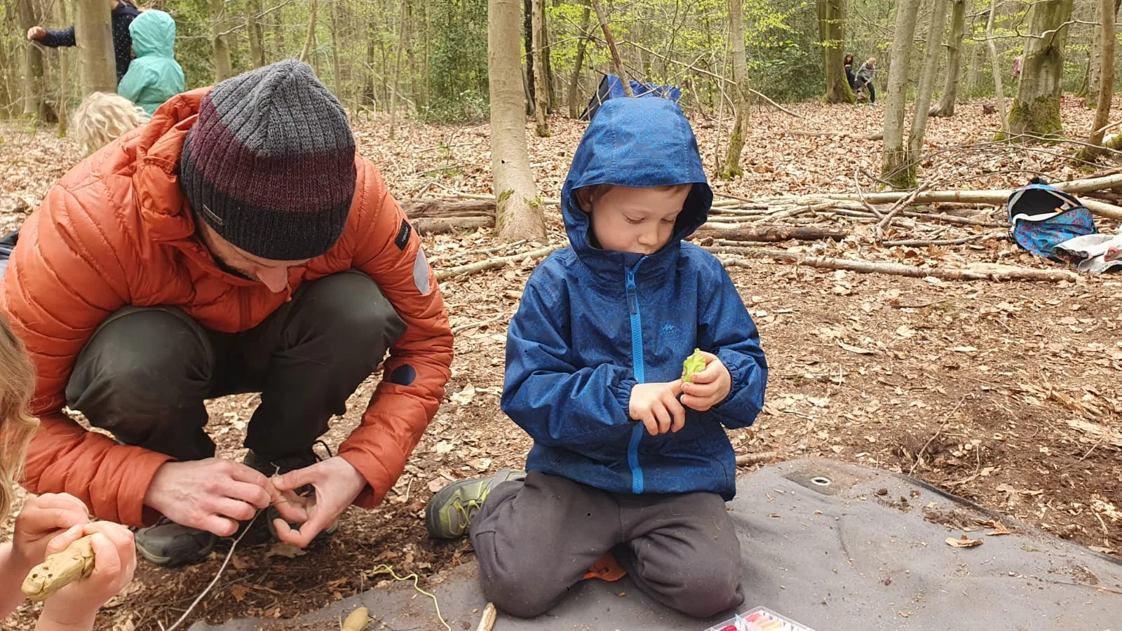 Un adulte et Léon, accroupis sur une bâche dans la forêt, en train de tailler du bois ensemble